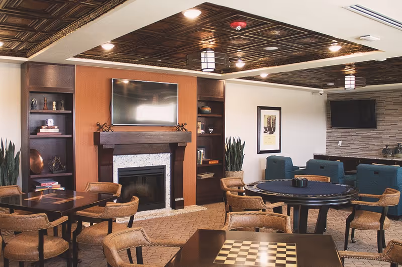 A warm communal lounge with a fireplace, flat-screen TVs, round and square game tables, and upholstered chairs.