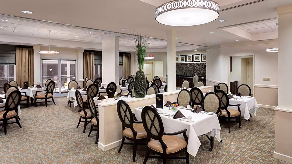 A spacious dining room with multiple tables covered in white tablecloths, each set with plates, cups, silverware, and folded brown napkins. The room features cushioned chairs with dark wooden frames, beige walls, carpeted floors, large windows with curtains, and modern circular ceiling lights. A tall decorative vase with green plants is placed on a half wall dividing the room.
