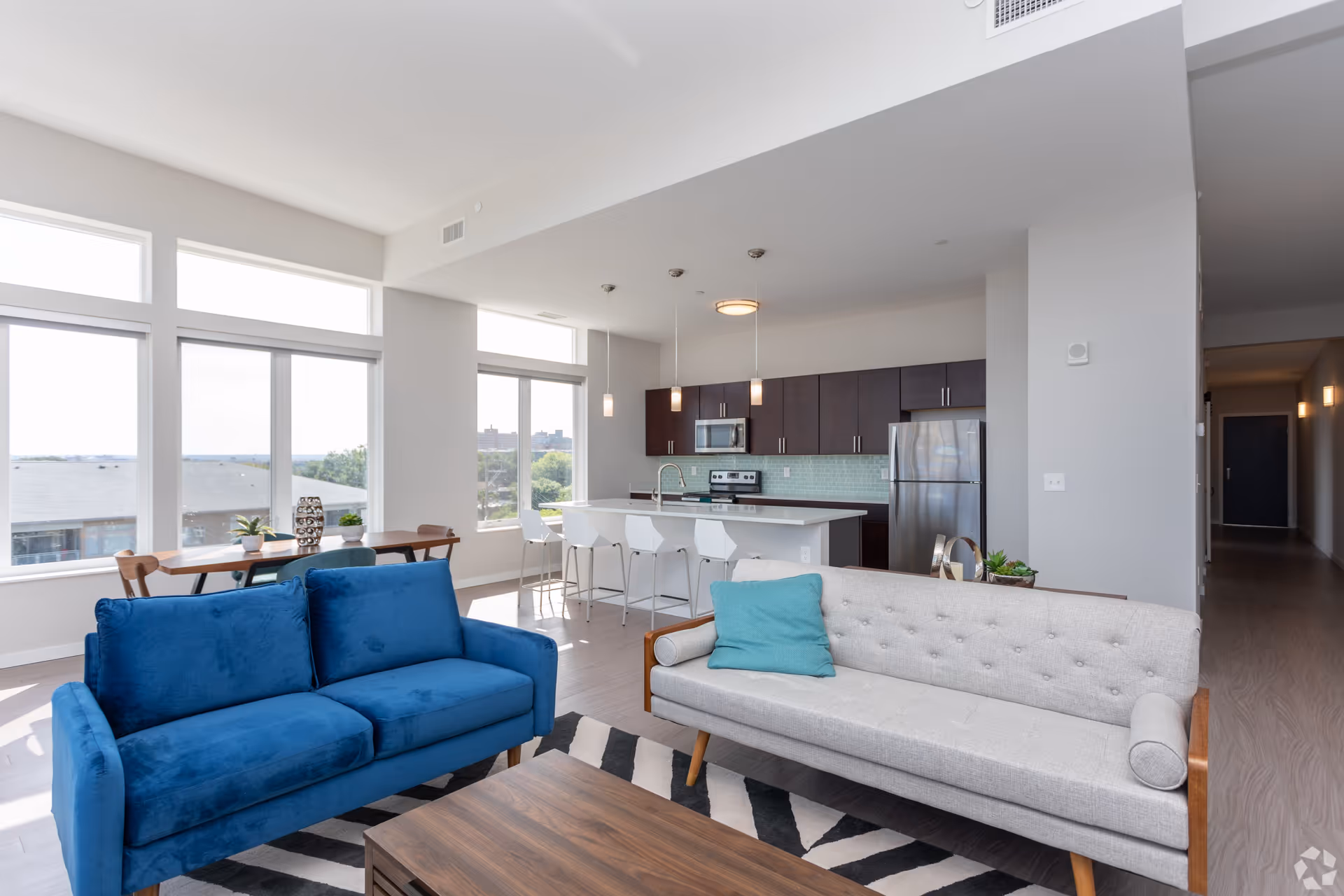 Bright and modern open living space featuring a blue sofa and a light gray tufted sofa with a teal pillow, a wooden coffee table on a black and white patterned rug, large windows letting in natural light, a dining table with chairs near the windows, and a kitchen area with dark wood cabinets, stainless steel appliances, a white island with four white bar stools, and pendant lights hanging above.