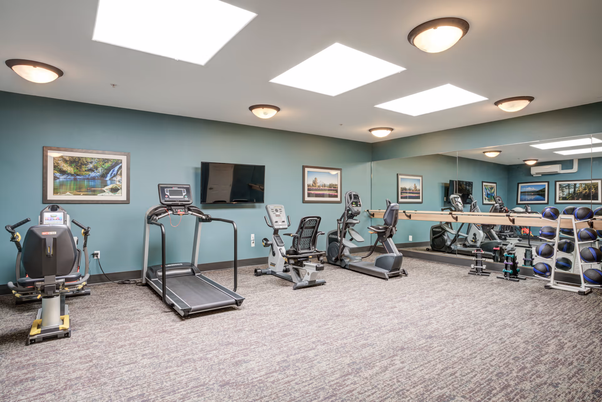 A senior living facility exercise room with various fitness equipment including a treadmill, recumbent bike, elliptical machine, and a rack of medicine balls. The room has teal walls, framed landscape pictures, a large wall mirror, ceiling lights, and skylights providing natural light.