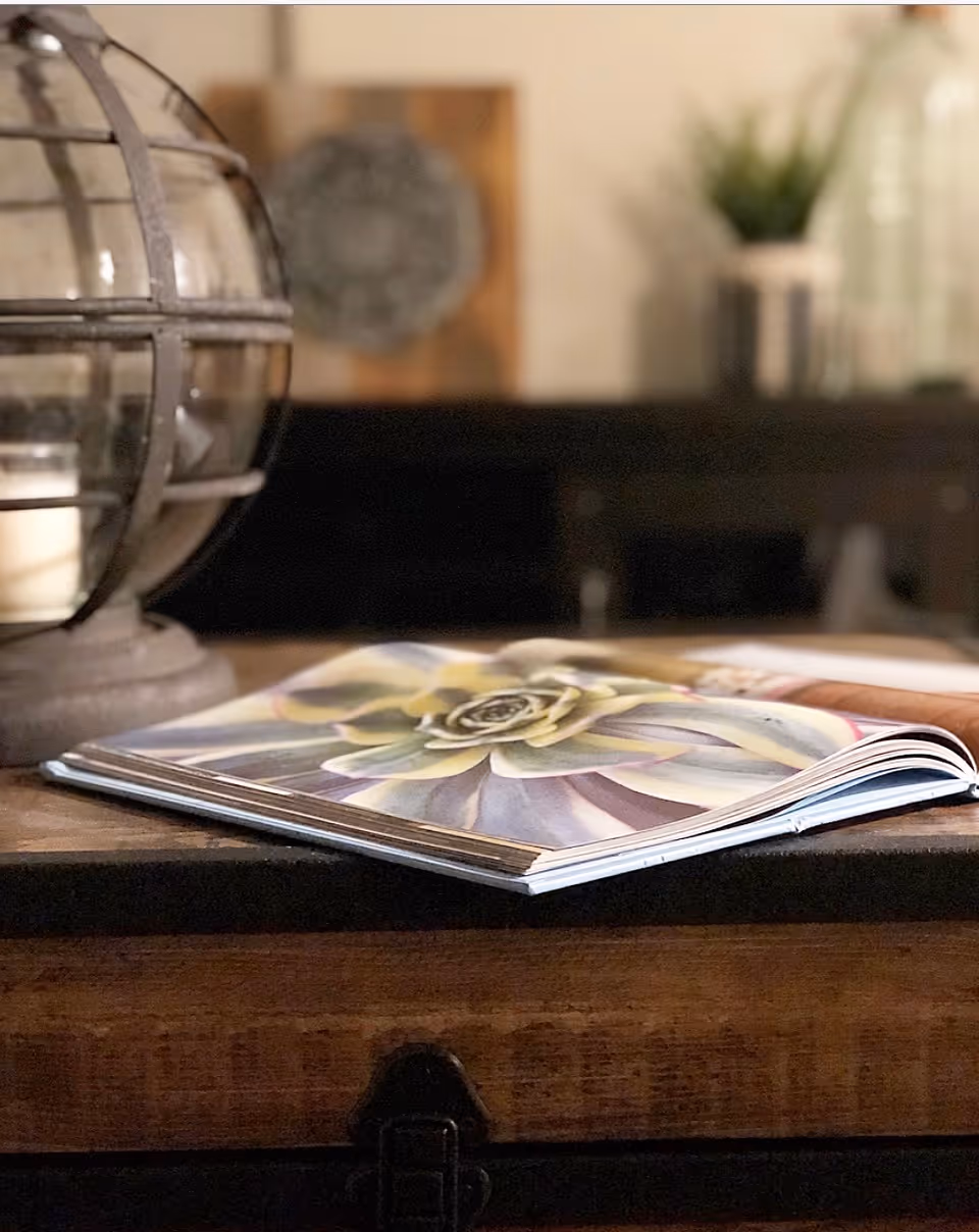 Close-up of an open book displaying a detailed image of a succulent plant, placed on a wooden surface with a decorative glass candle holder nearby and blurred background featuring plants and decor.