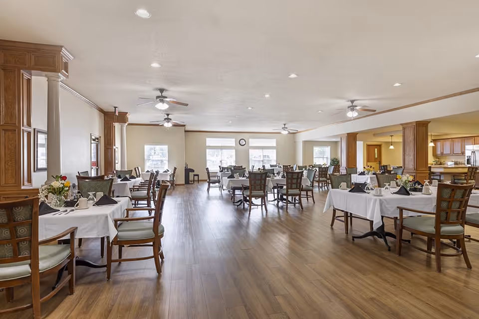 A spacious dining room with multiple tables covered in white tablecloths, each set with black napkins, cups, and flower arrangements. The room has wooden flooring, ceiling fans, and large windows letting in natural light. The background includes a kitchen area with wooden cabinetry.