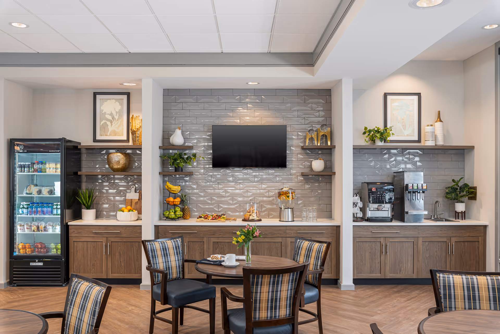 A modern common area with a refreshment station featuring a refrigerator stocked with drinks and snacks, a counter with fruit and a beverage dispenser, a coffee machine, and a soda fountain. The wall behind the counters is decorated with gray subway tiles and shelves holding decorative items and plants. In front, there are round tables with plaid cushioned chairs, and a flat-screen TV mounted on the wall.