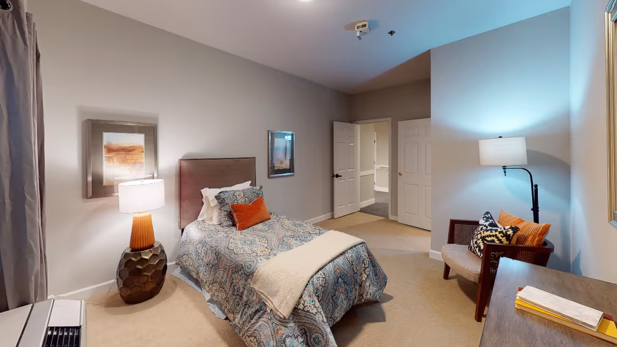 A furnished single bedroom with a twin bed, patterned comforter, bedside lamp, armchair, and an open door to a hallway.