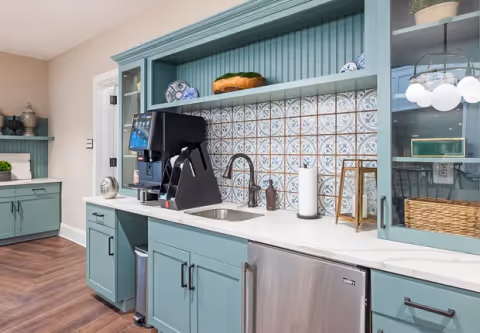A compact kitchenette with teal cabinets, patterned tile backsplash, a coffee machine, sink, and stainless mini-fridge.