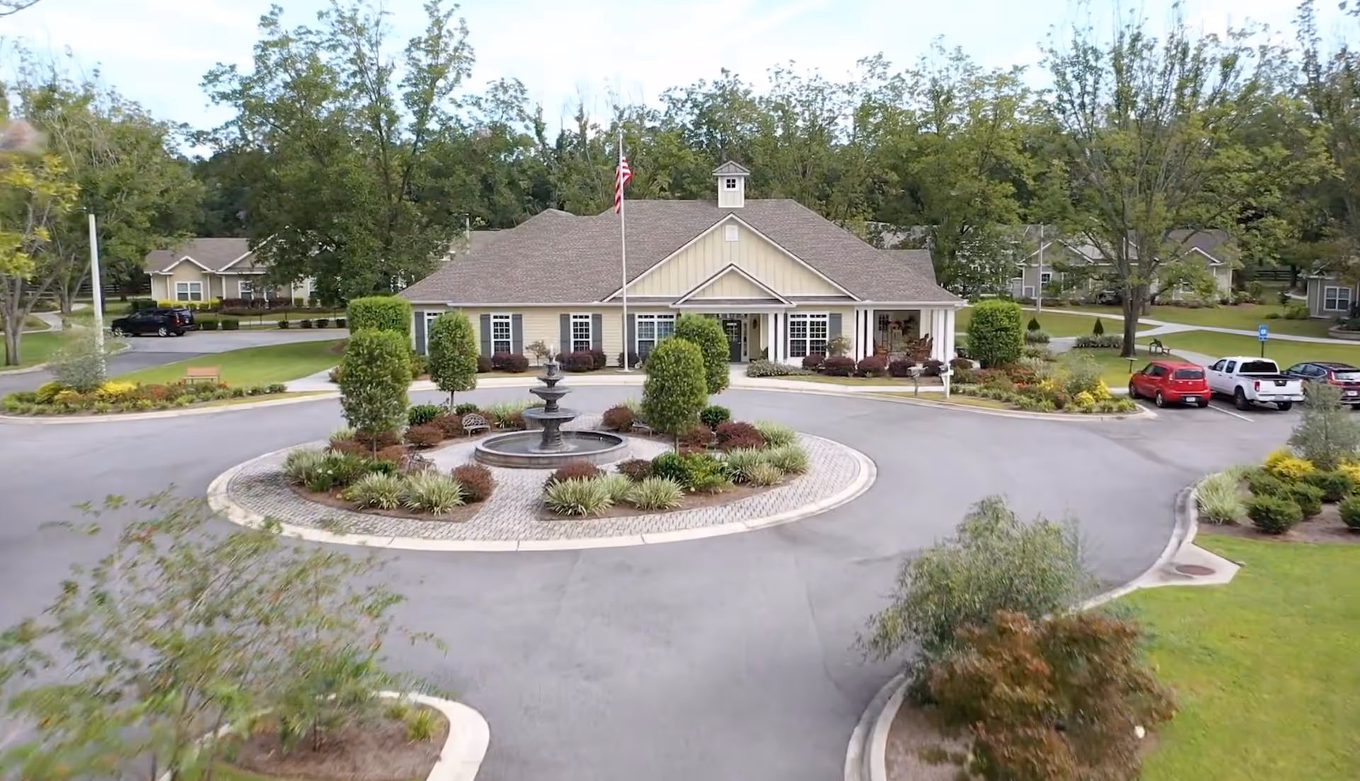 Front exterior view of a single-story building with a pitched roof and a central cupola, surrounded by landscaped greenery and a circular driveway with a fountain in the center. Several cars are parked on the right side, and trees and shrubs are visible around the property.