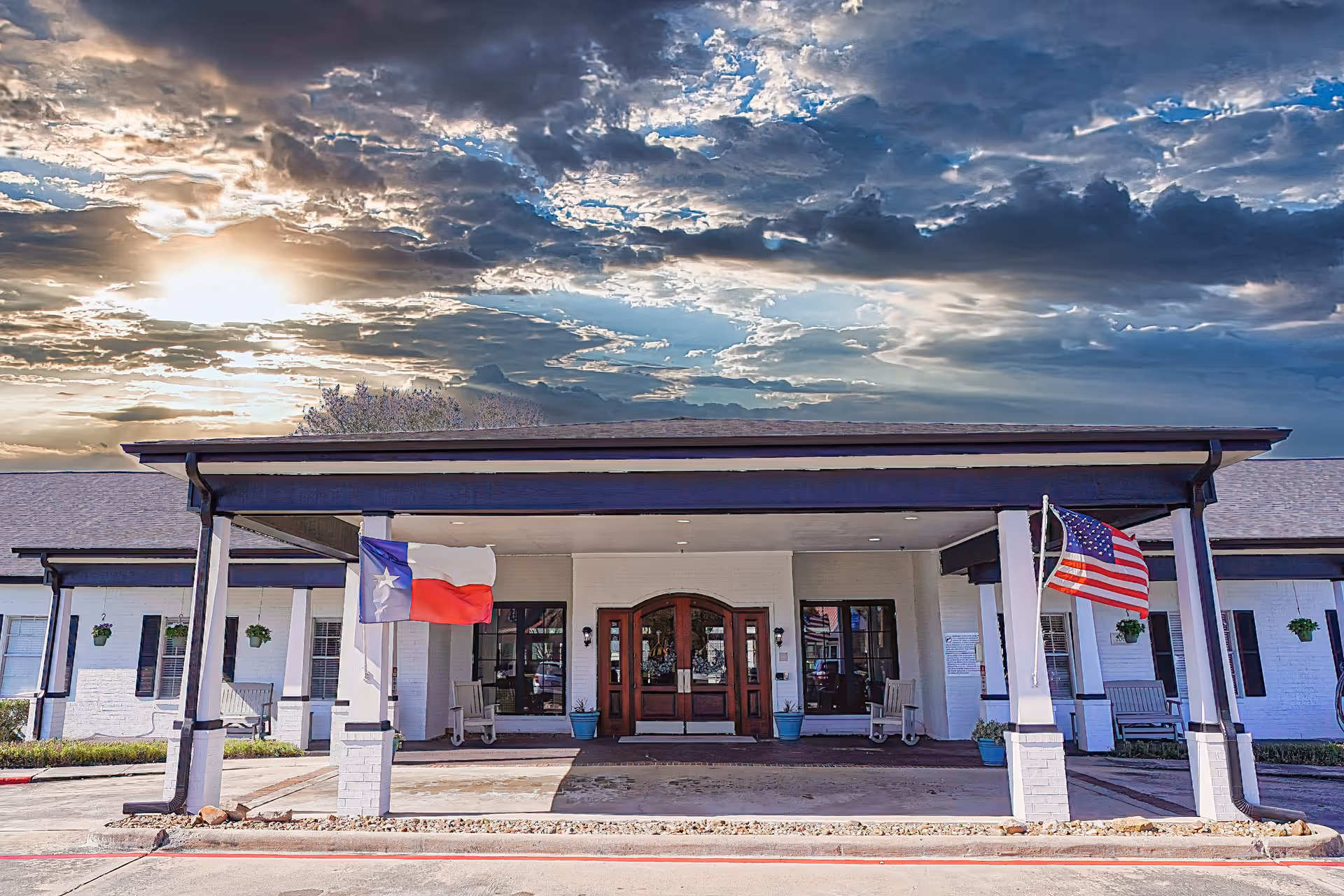 Front exterior view of Sodalis Lake Jackson facility with a covered entrance, two flags (Texas state flag and American flag) on either side, rocking chairs, potted plants, and a dramatic cloudy sky at sunset.