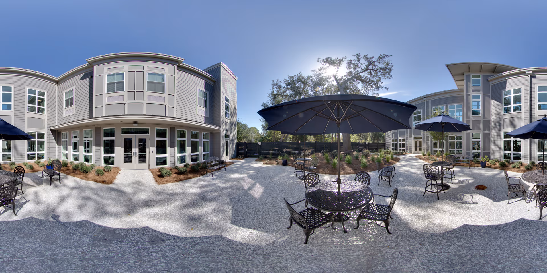 Outdoor courtyard area of Indigo Hall Assisted Living and Memory Care featuring multiple round metal tables with chairs and large blue umbrellas. The courtyard is surrounded by a two-story building with many windows and landscaped with small plants and mulch beds under a clear blue sky.