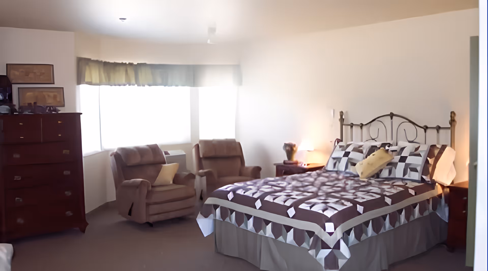 A bedroom with a bed covered in a patterned quilt and multiple pillows, two cushioned armchairs with a small table between them near a window with curtains, and a wooden dresser with framed pictures on top.