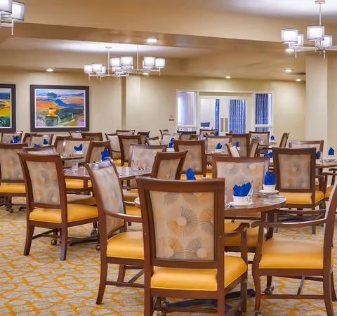 Spacious dining room with round tables and wooden chairs set with place settings and blue napkins.