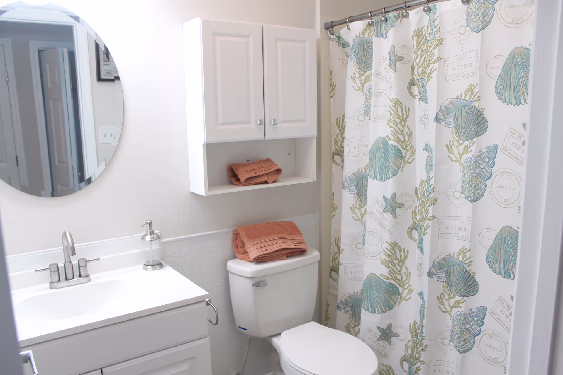 A clean bathroom featuring a white vanity with a silver faucet, a round mirror above the sink, a white toilet with folded pink towels on top, a white wall-mounted cabinet with additional pink towels, and a shower with a curtain decorated with seashell and starfish patterns.