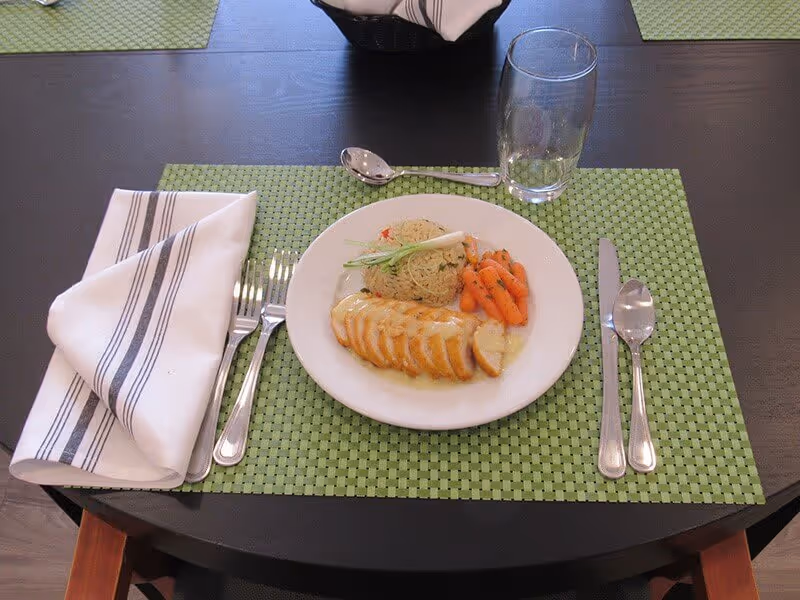 A dining table set with a green woven placemat, a white plate containing sliced chicken breast with sauce, a serving of rice, and cooked baby carrots. The table setting includes a folded white napkin with black stripes, a fork and knife on the left, a spoon and knife on the right, a spoon above the plate, and an empty drinking glass to the upper right of the plate.