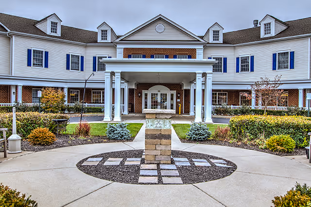 Front exterior view of a two-story senior living facility with white siding, blue shutters, and a covered entrance supported by white columns. There is a circular driveway with landscaped bushes and a decorative stone pillar in the center.
