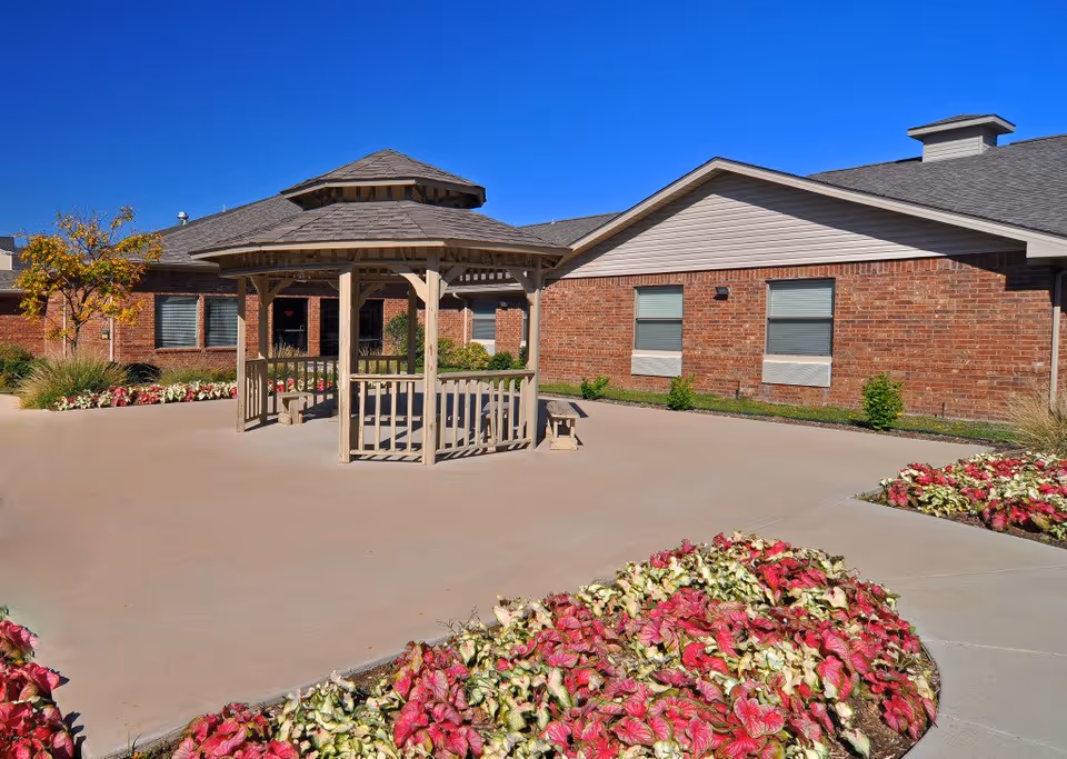 Outdoor courtyard area with a wooden gazebo in the center surrounded by flower beds with red and white flowers. The background shows a brick building with several windows under a clear blue sky.