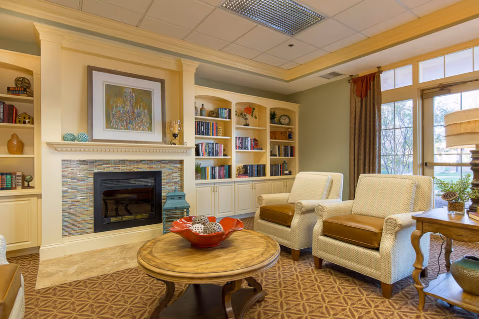 A cozy living room area featuring two cushioned armchairs with brown seat cushions, a round wooden coffee table with a red decorative bowl filled with ornamental balls, built-in cream-colored bookshelves filled with books and decorative items, a fireplace with a mosaic tile surround, and a large window with curtains letting in natural light.