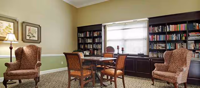 Sunlit common room with built-in bookshelves around a window, a central table with four chairs and two upholstered armchairs.