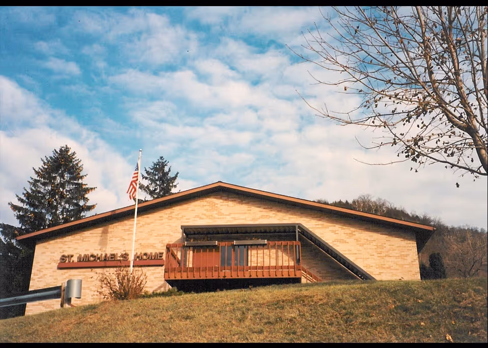Exterior view of St. Michael's Home building with a brick facade, a small balcony, an American flag on a flagpole, and a grassy hill in the foreground under a partly cloudy sky.