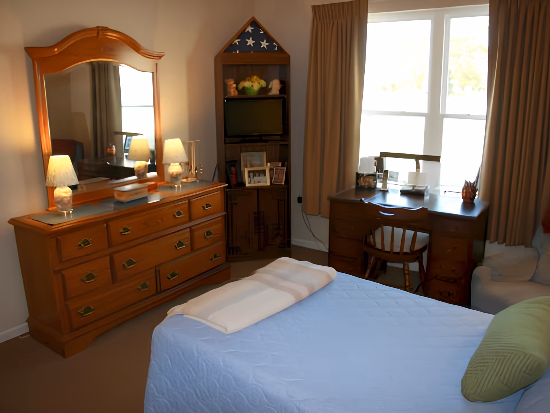 A cozy bedroom with a wooden dresser topped with two lamps and a large mirror, a bed with a light blue quilt and a green pillow, a wooden desk with a chair near a window with beige curtains, and a corner shelf holding a folded American flag, a TV, and framed photos.