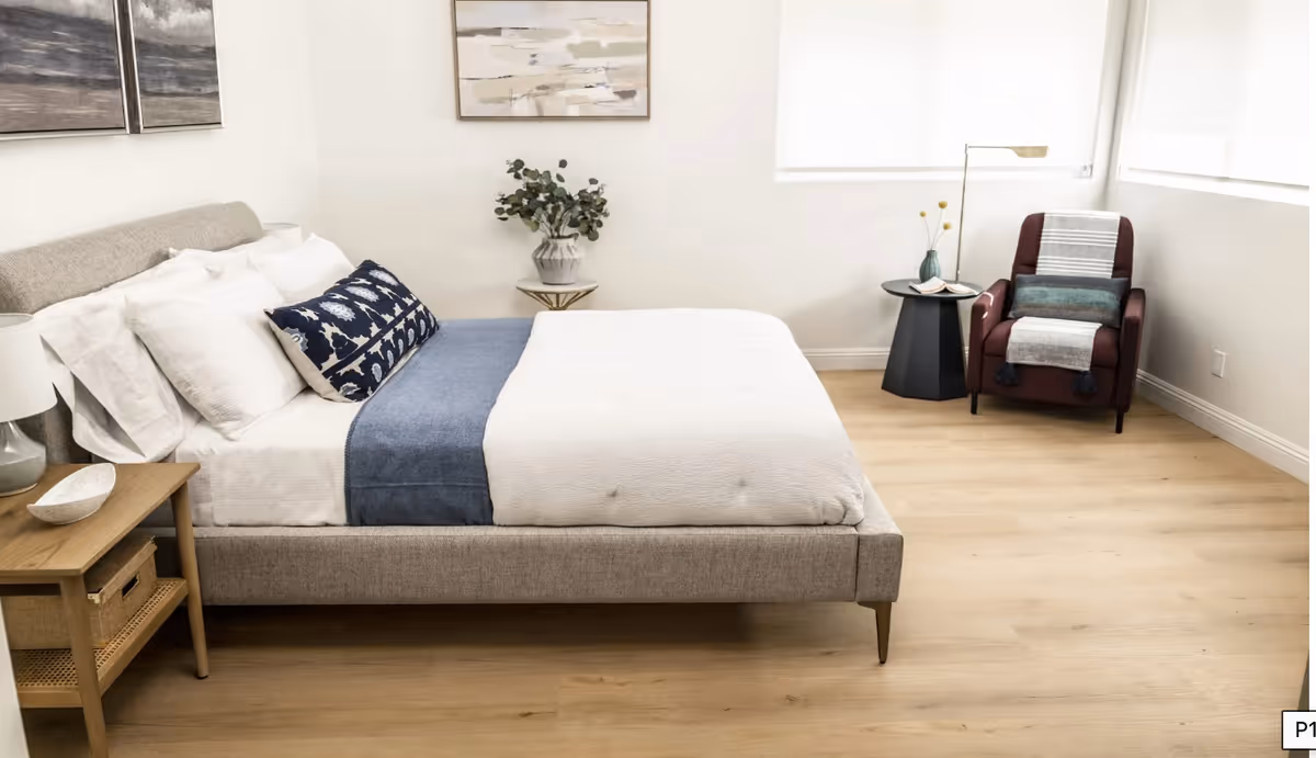 Bright minimalist bedroom with a gray upholstered bed dressed in white and blue linens, a wooden nightstand, and a chair by the window.