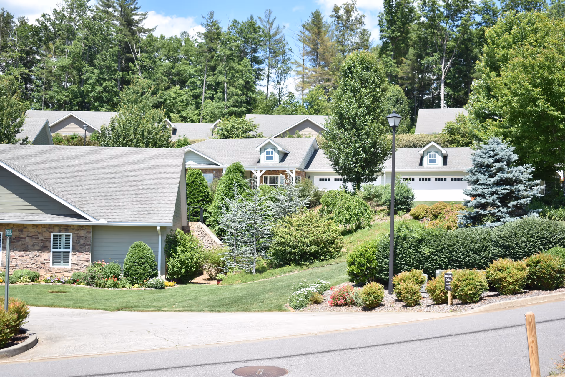Manicured hillside of single-story residential buildings with garages, shrubs, lawns, and tall trees under a blue sky.