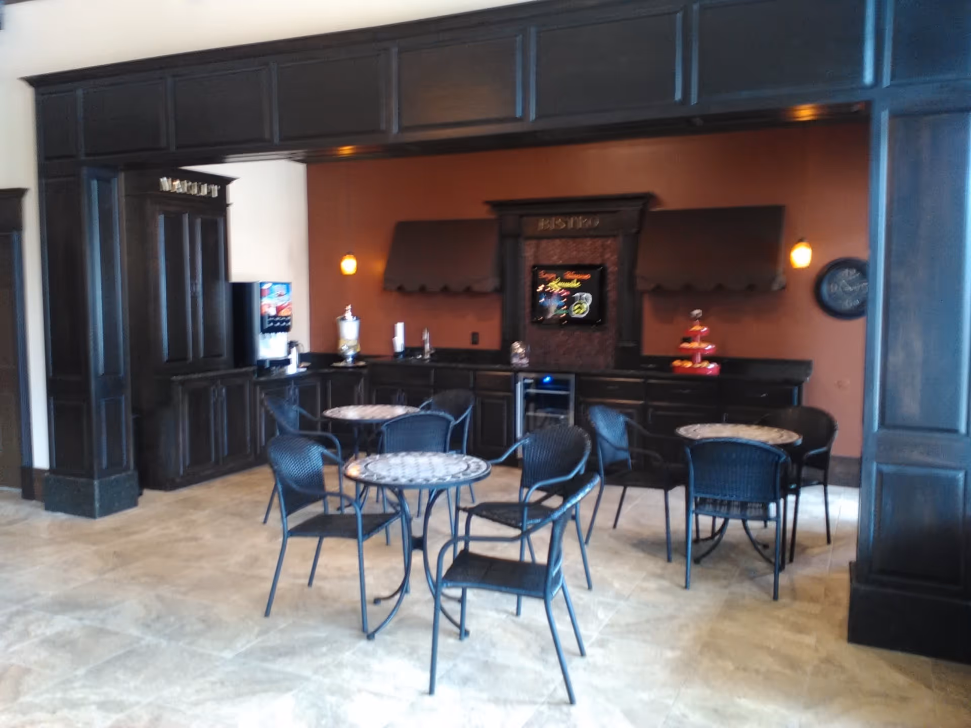 A cozy bistro area with three round mosaic-tiled tables and black metal chairs. The back wall features dark wood cabinetry with a small beverage dispenser, a water dispenser, and a tiered tray with snacks. The wall is painted a warm reddish-brown color with two small pendant lights and a decorative sign that reads 'BISTRO'.