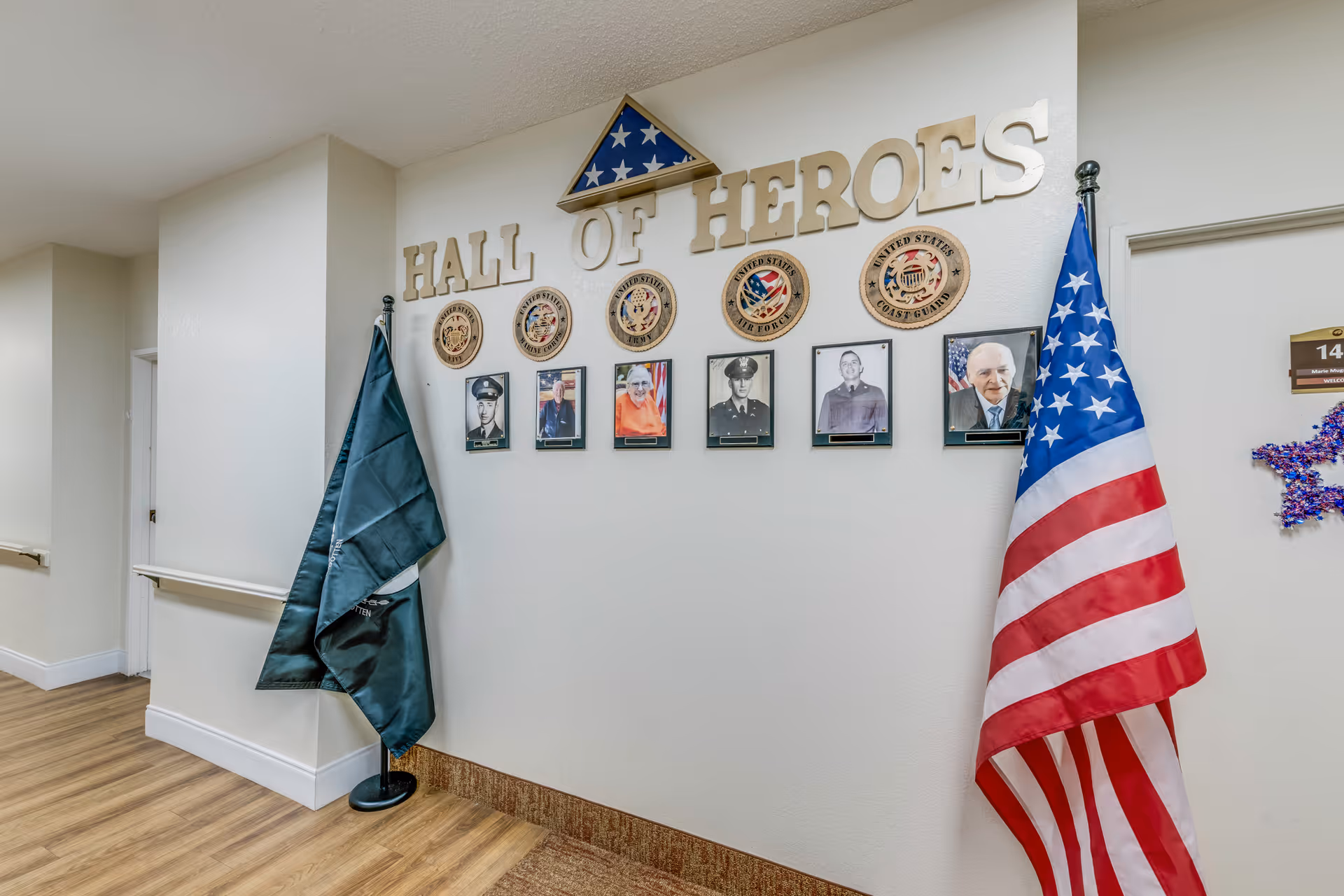 Interior hallway wall display titled 'Hall of Heroes' featuring plaques and framed photos of veterans from different branches of the United States military, with an American flag and another flag on stands on either side.