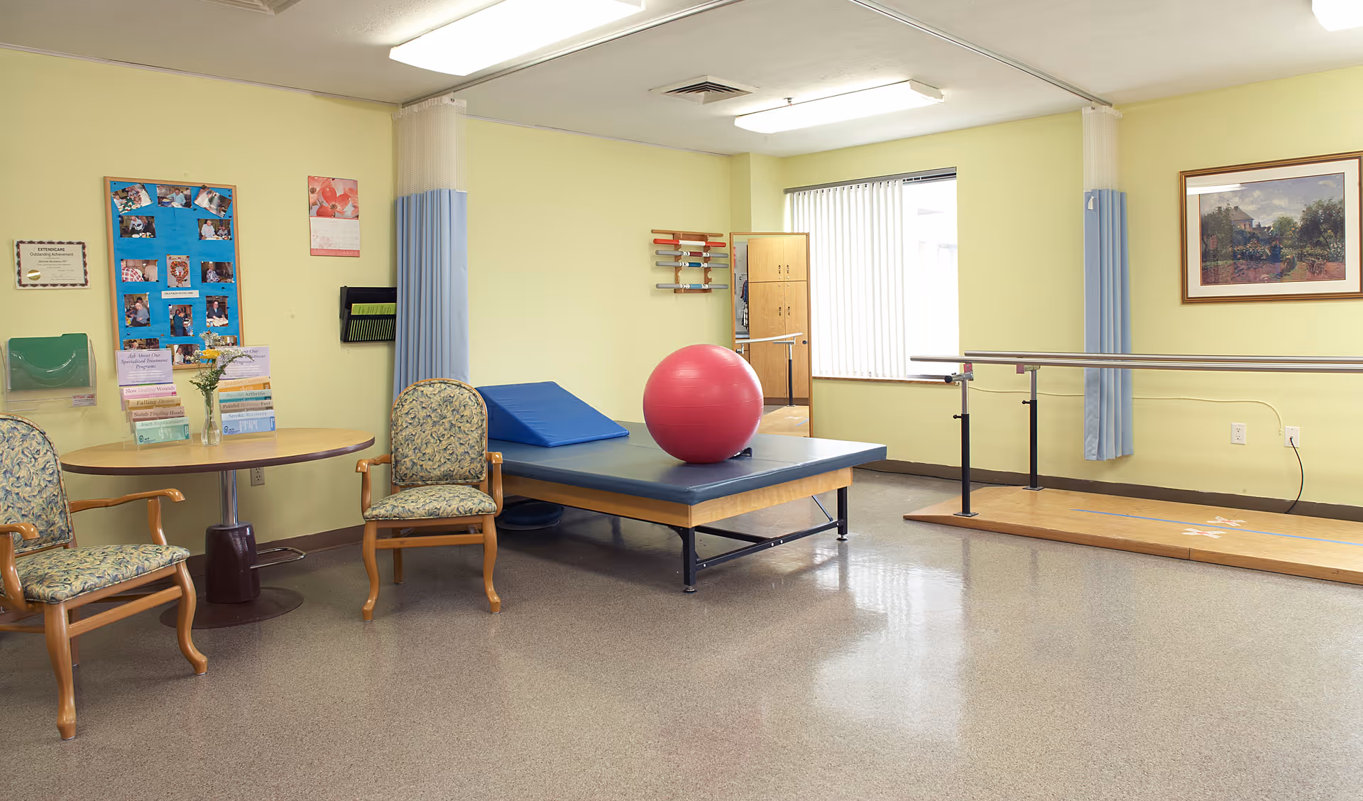 A bright interior therapy room with a padded treatment table, a large pink exercise ball, parallel bars, a small table with chairs, and bulletin boards on the wall.