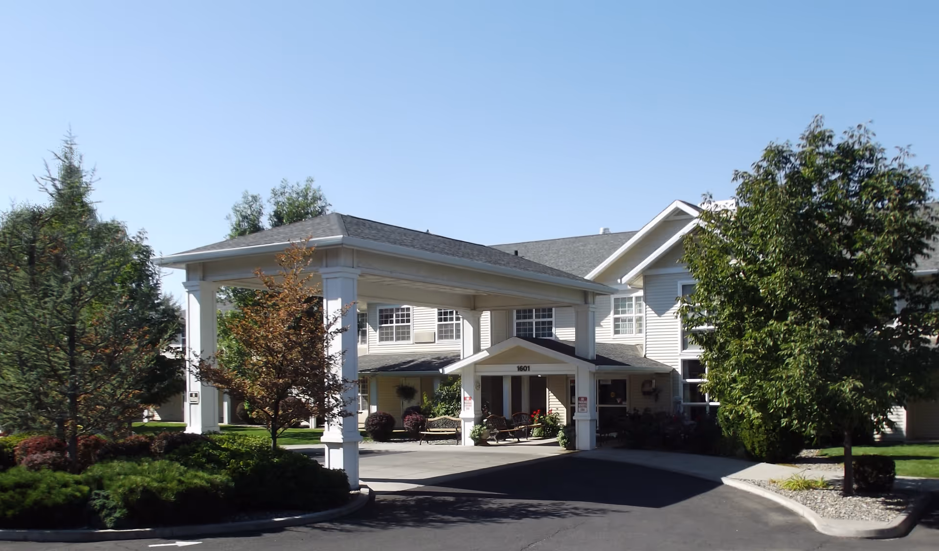 Exterior view of McKay Creek Assisted Living facility showing the main entrance with a covered drop-off area, surrounded by trees and landscaped greenery under a clear blue sky.