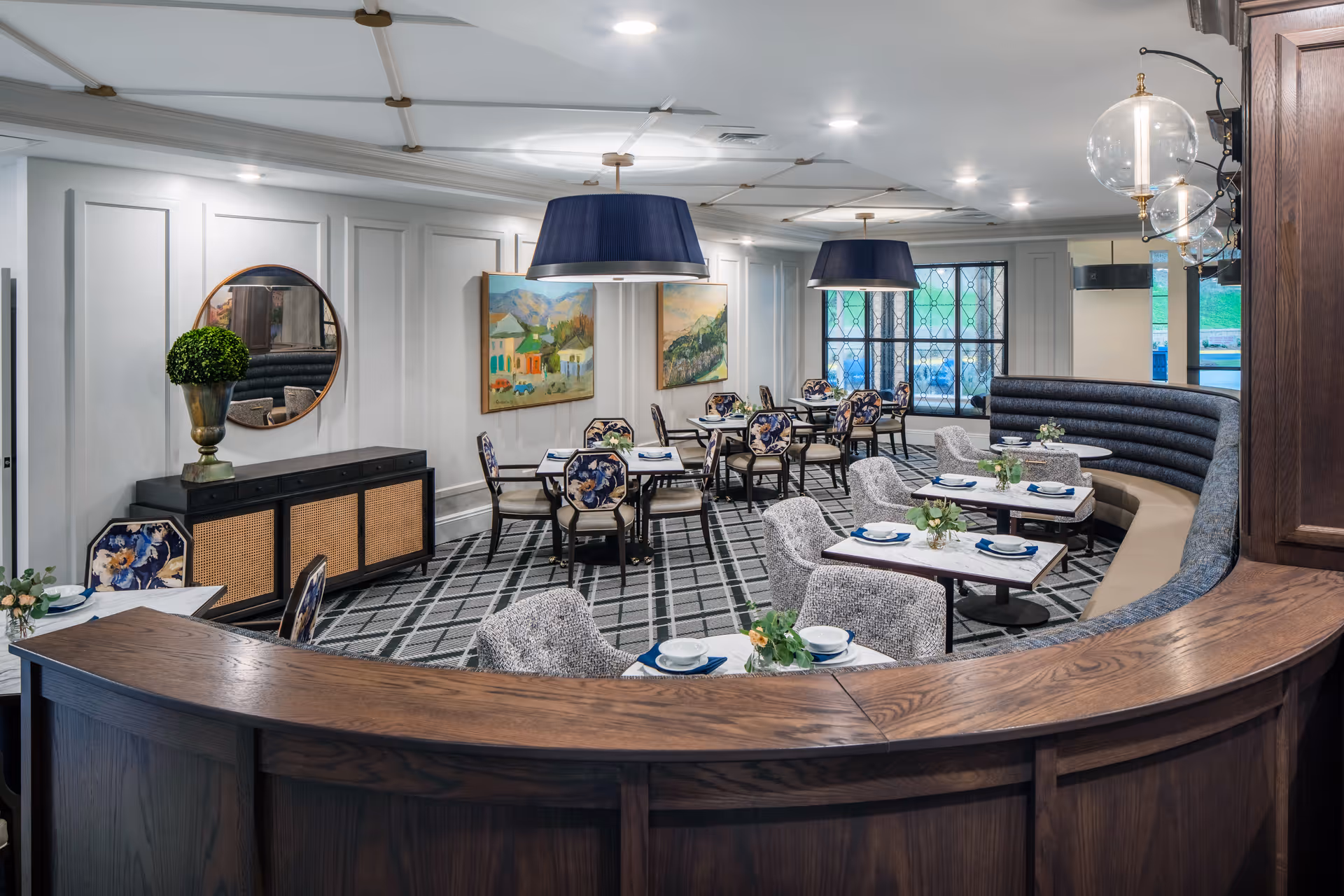 Well-appointed dining room with a curved wooden counter, round tables set with dishes, upholstered chairs and banquette seating under pendant lights.
