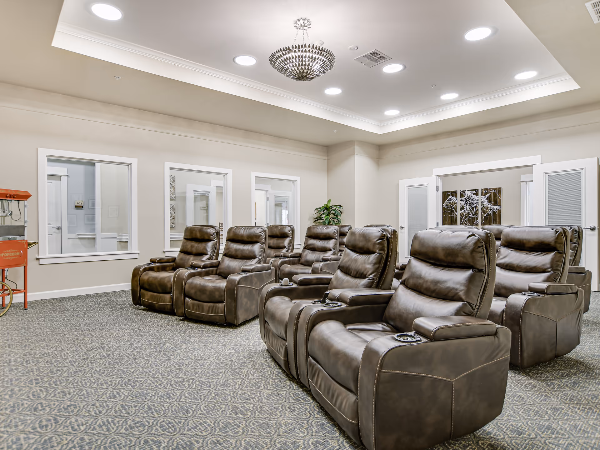 A spacious room with two rows of brown leather recliner chairs arranged facing forward. The room has a patterned carpet, beige walls, and a decorative ceiling light fixture. There are windows and doors along the walls, and a red popcorn machine is visible on the left side.