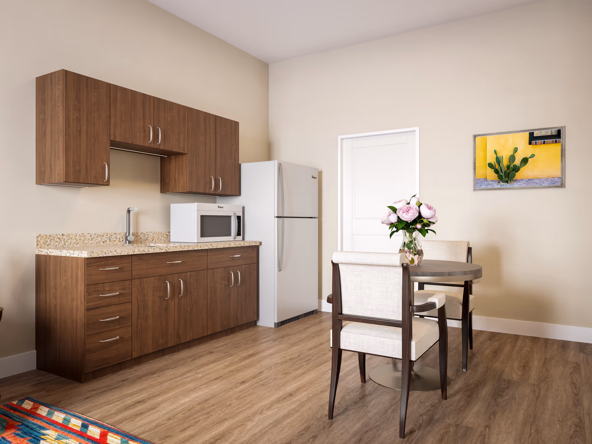 A small kitchenette with wood cabinets, a refrigerator and microwave, and a round dining table with chairs and a vase of flowers.