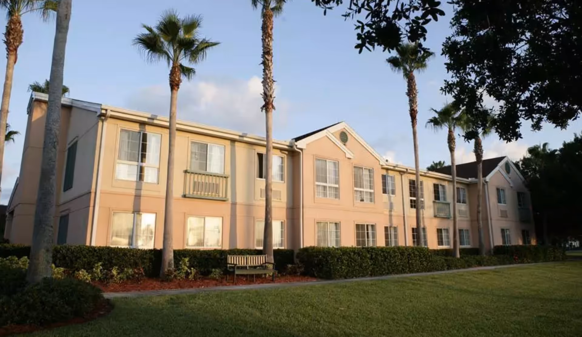 Exterior view of a two-story senior living facility building with beige and light peach walls, multiple windows, palm trees, green bushes, a bench, and a well-maintained lawn under a clear sky.