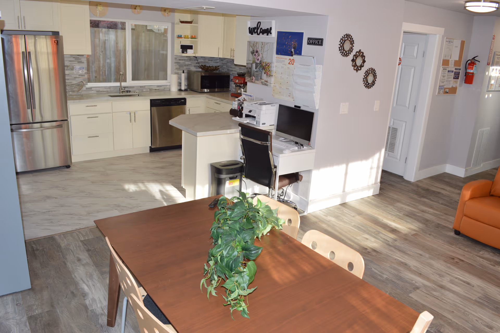 Interior view of a senior living facility showing a modern kitchen with white cabinets, stainless steel refrigerator and dishwasher, a small office desk with a computer and printer, a wooden dining table with chairs and a green plant centerpiece, and part of an orange armchair in the living area.