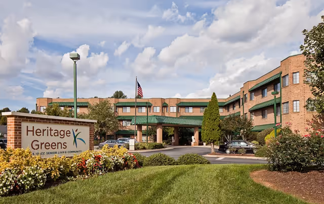 Brick multi-story senior living building with green awnings, an entrance canopy and flagpole, and a landscaped lawn with a 'Heritage Greens' sign in the foreground.