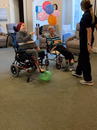 Two elderly women in wheelchairs playing with colorful balloons indoors while a caregiver stands nearby in a living room setting with armchairs and large windows.