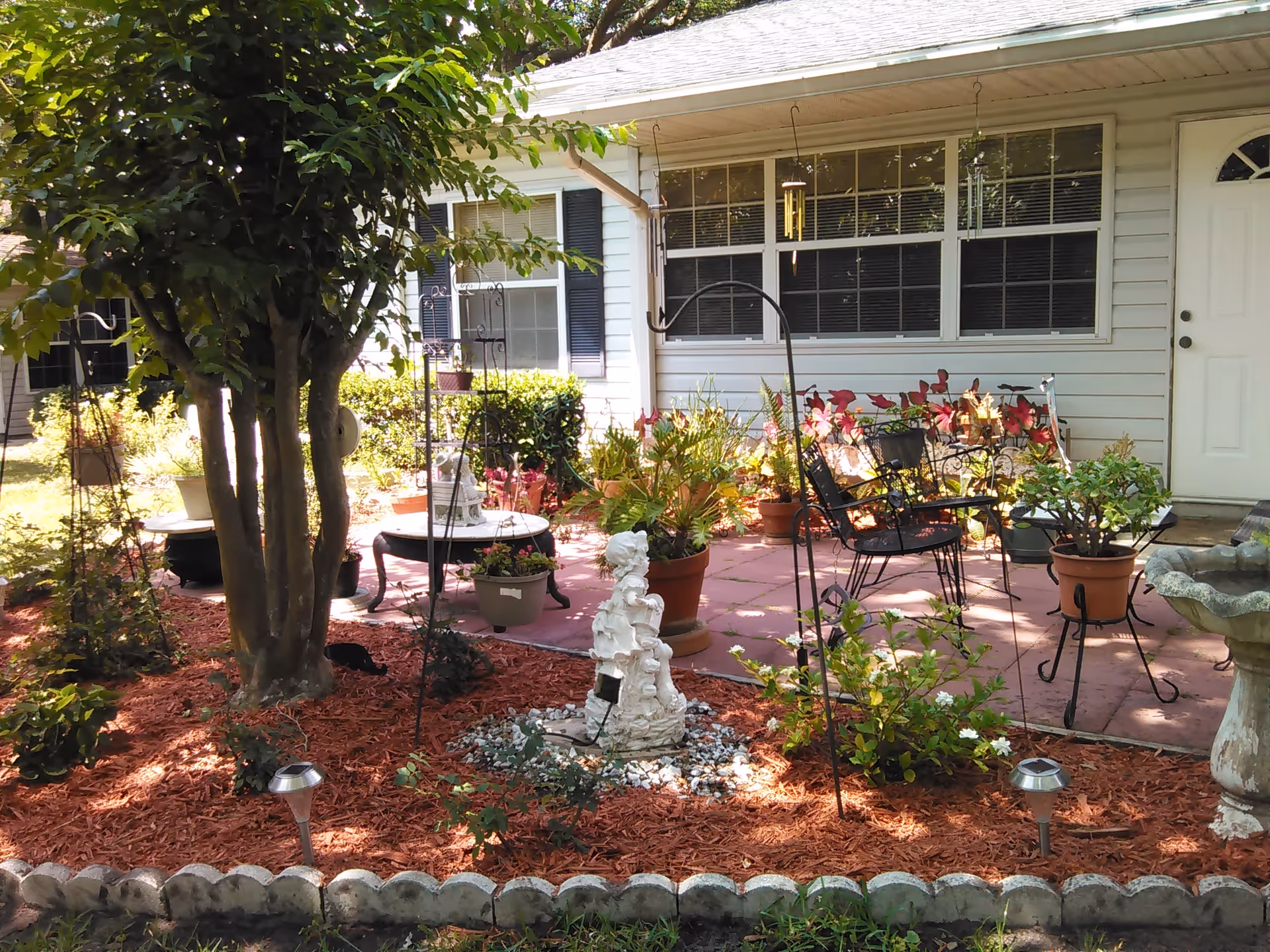 A peaceful outdoor garden area with a variety of potted plants, a small white statue, garden decorations, and outdoor seating including a metal bench and chairs on a paved patio in front of a white building with windows and a door.