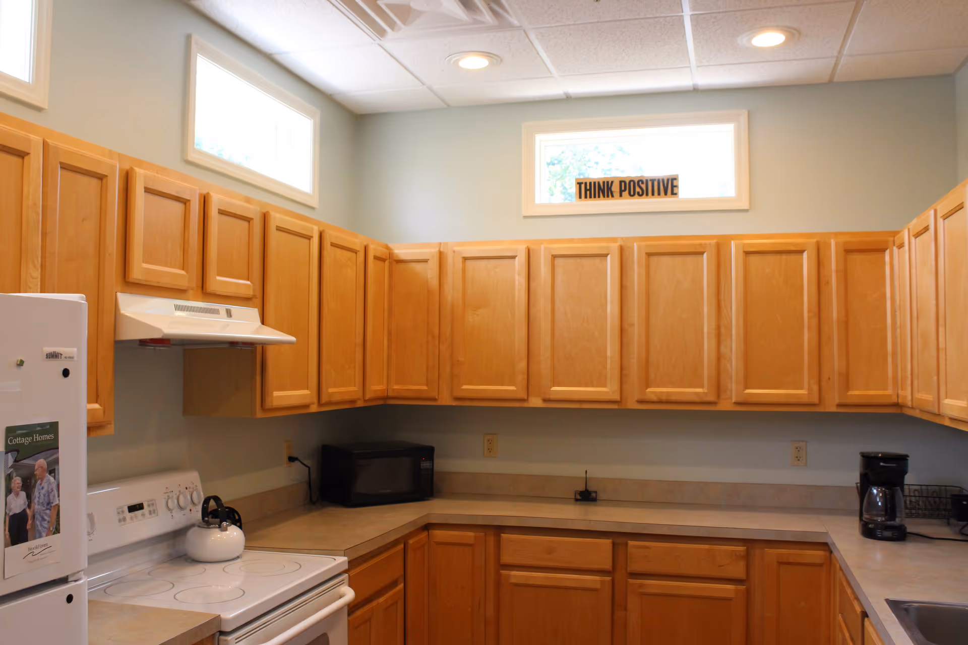 A kitchen with light wood cabinets, a white stove with a kettle on it, a microwave, a coffee maker, and a refrigerator. There are two small windows near the ceiling, and a sign between the windows reads 'THINK POSITIVE'.