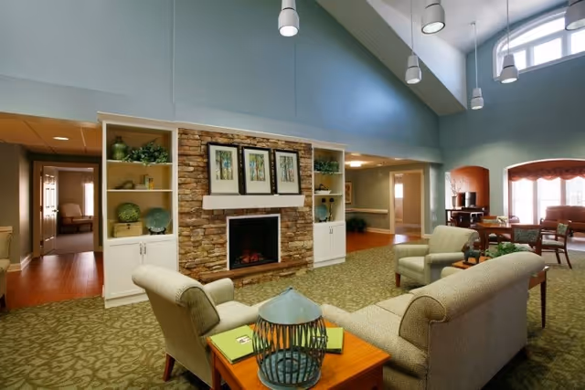 A spacious living room area in a senior living facility with high ceilings and large windows. The room features a stone fireplace with three framed pictures above it, flanked by built-in white shelves decorated with plants and ornaments. Comfortable upholstered chairs and sofas are arranged around a wooden coffee table with decorative items. In the background, there is a dining area with tables and chairs near a window with a red valance.