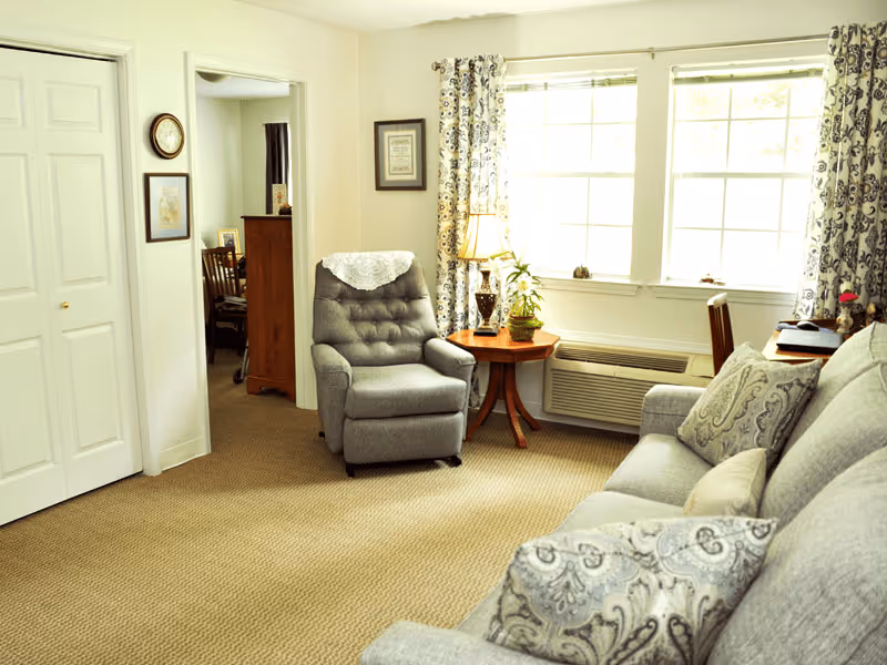 A cozy living room in an assisted living facility featuring a gray cushioned armchair with a white doily on the headrest, a matching gray sofa with patterned pillows, a wooden side table with a lamp and a plant, large windows with patterned curtains letting in natural light, beige carpeted floor, and a glimpse into an adjacent room with wooden furniture.