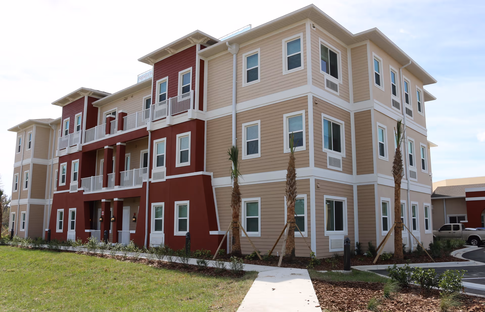 Exterior view of a three-story senior living facility building with beige and red siding, multiple windows, balconies, and a small landscaped area with young palm trees and a sidewalk leading to the entrance.