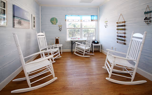 Bright small sitting room with four white wooden rocking chairs on a wood floor facing a window.