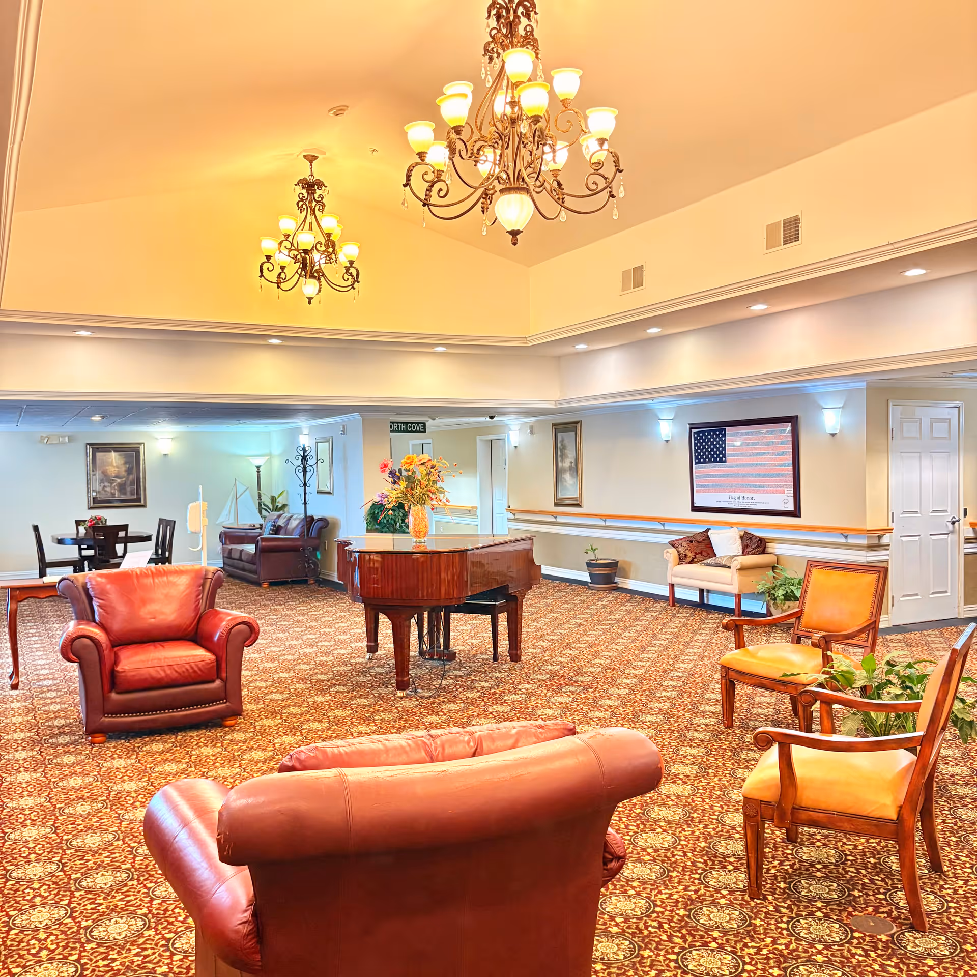A spacious and elegantly decorated common area in an assisted living facility featuring a grand piano with a floral arrangement on top, multiple comfortable leather armchairs, wooden chairs with cushions, and a patterned carpet. The room is illuminated by ornate chandeliers hanging from a high ceiling. There are framed pictures and plants along the walls, and a door is visible on the right side.