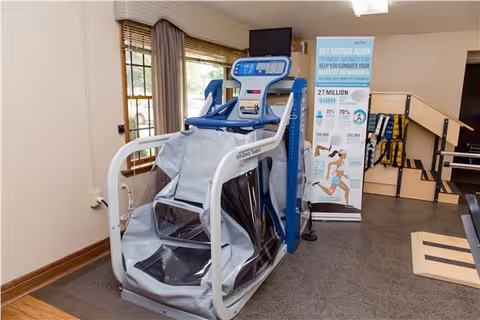 Physical therapy room with a large exercise machine designed for assisted walking or movement therapy. A window with blinds and curtains is visible on the left wall. Behind the machine, there is a standing informational banner about physical therapy benefits and statistics. The room has a brown floor and light-colored walls.