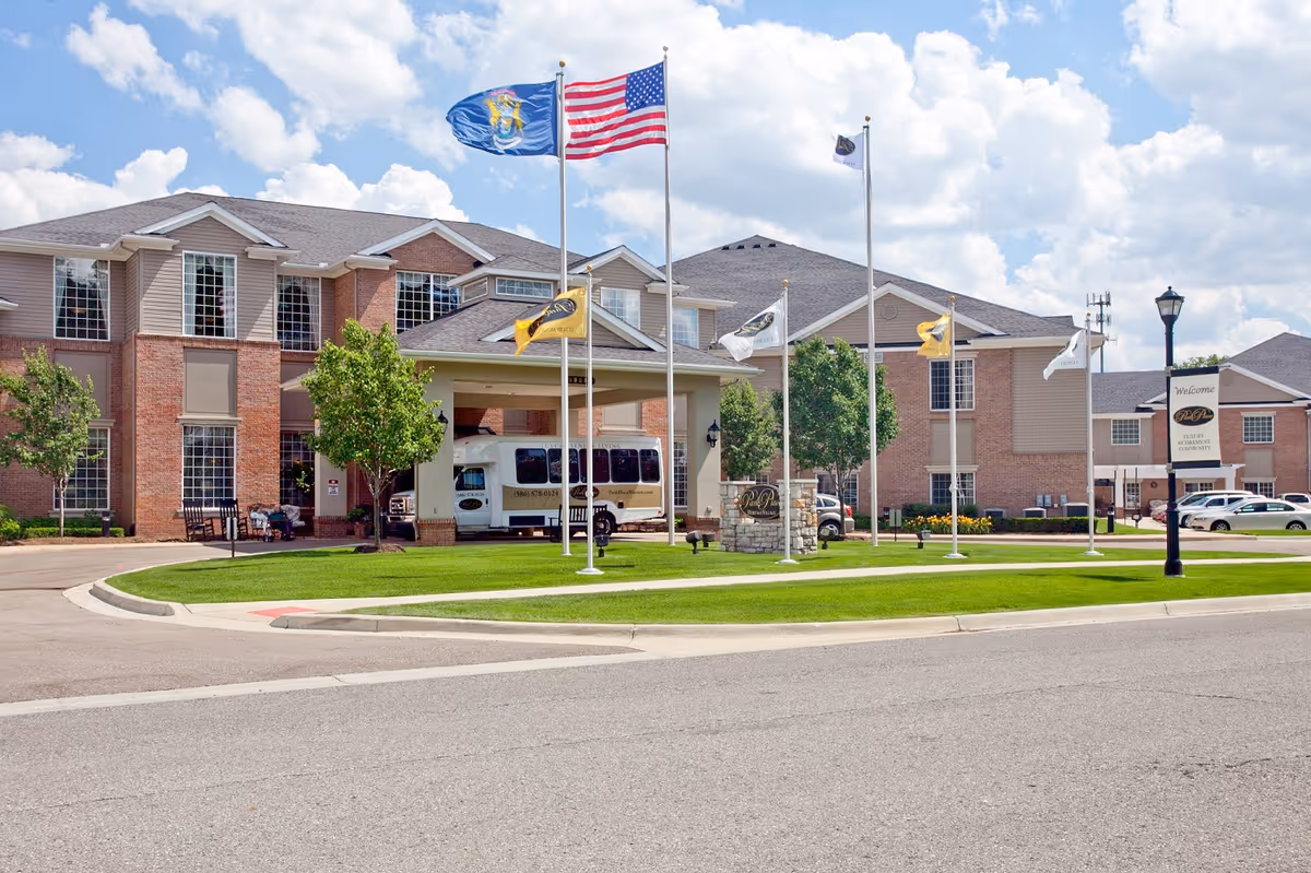 Exterior view of a senior living facility with a brick and beige siding building, several flagpoles with flags including the American flag, a driveway, green lawn, and a shuttle bus parked under a covered entrance.