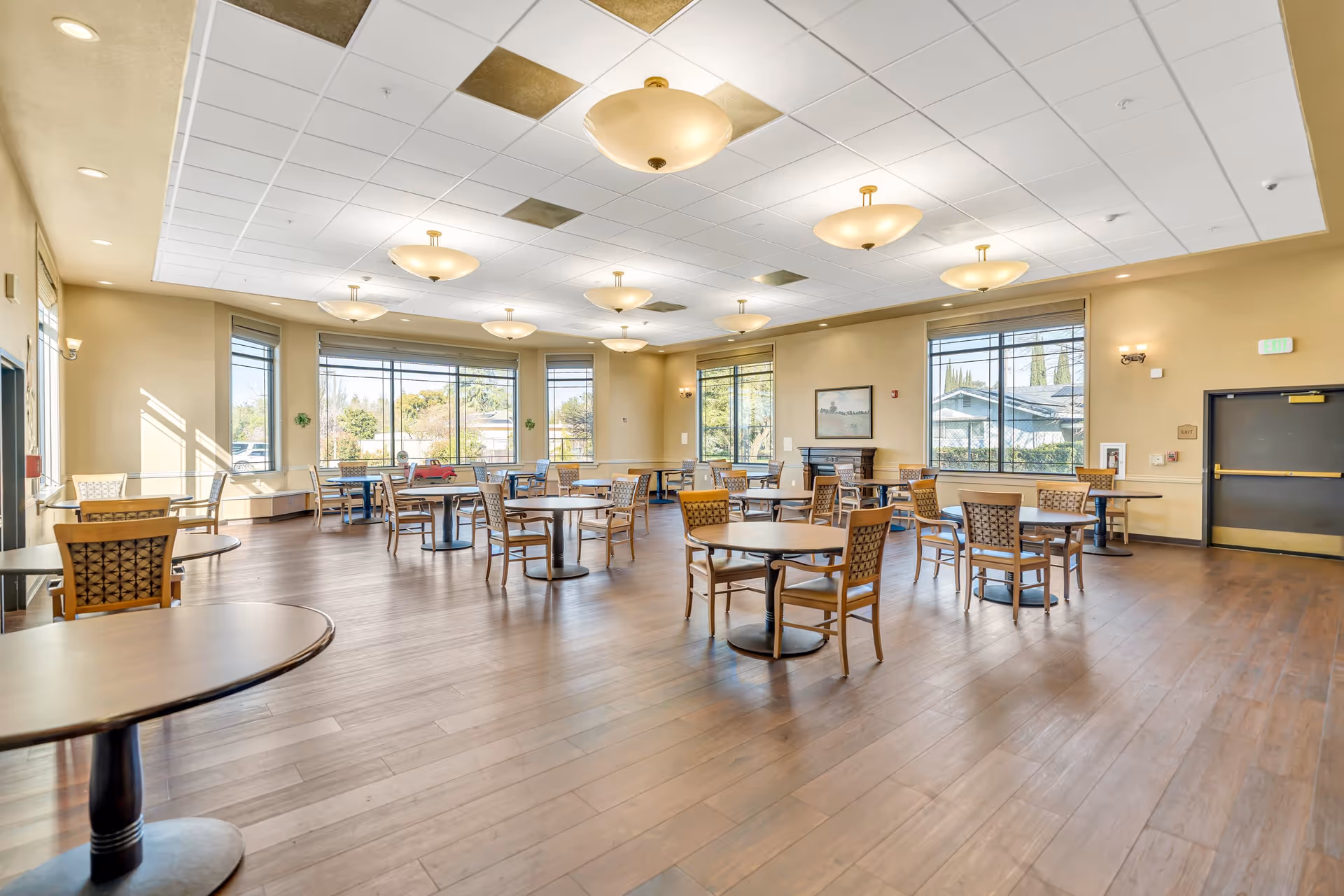 A spacious dining room with multiple round tables and chairs arranged neatly on a wooden floor. Large windows allow natural light to fill the room, and ceiling lights provide additional illumination. The walls are painted a warm beige color, and there is a door on the right side of the room.