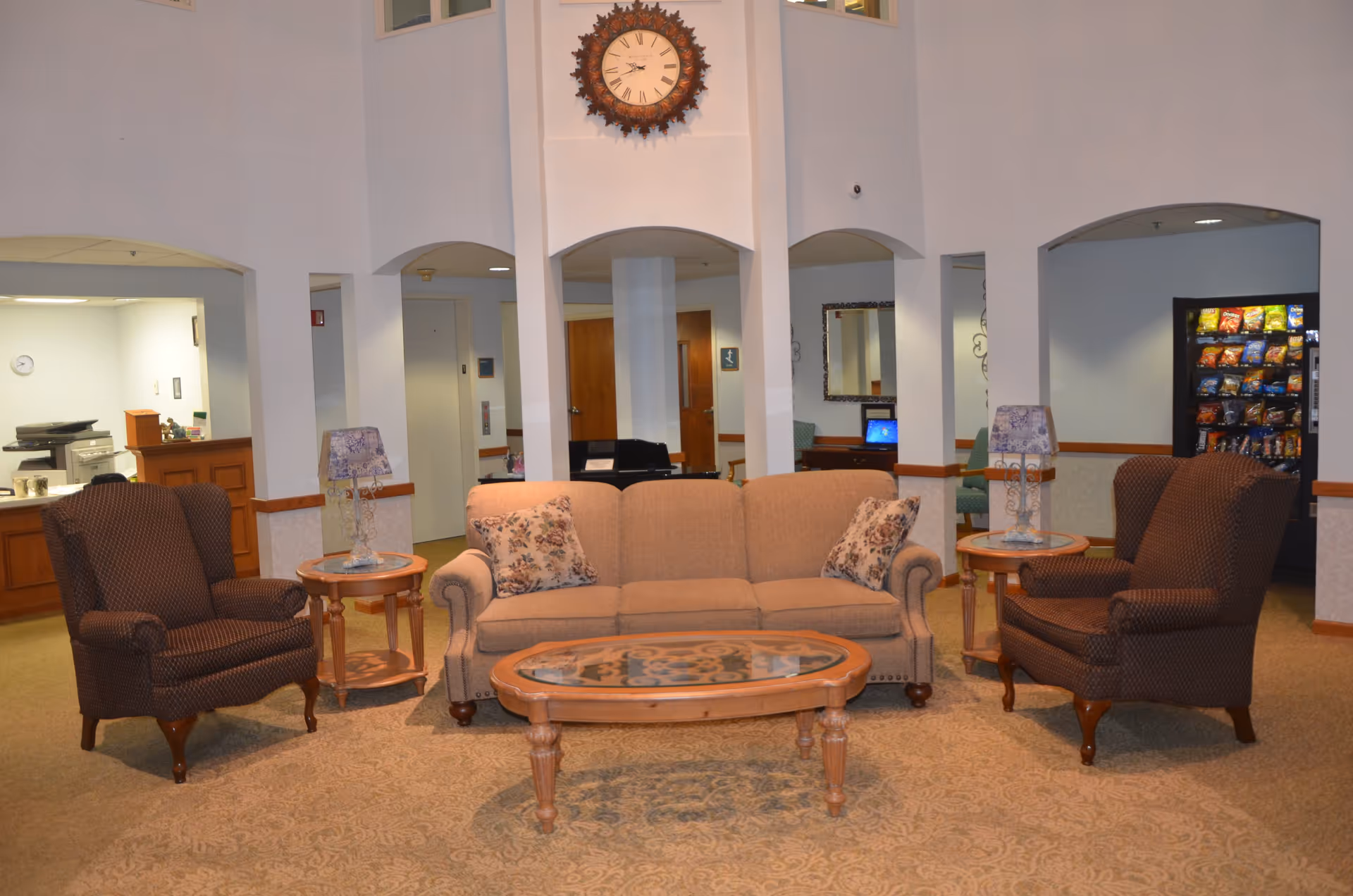 A cozy seating area in a senior living facility with a beige sofa adorned with floral cushions, two brown patterned armchairs, and a wooden coffee table with a glass top. Two side tables with decorative lamps flank the sofa. In the background, there is a vending machine, a reception desk, and a wall clock with a decorative frame above the seating area.