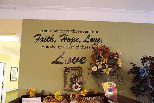 Interior wall of a senior living facility with a decorative quote that reads 'And now these three remain: Faith, Hope, Love, But the greatest of these is Love'. Below the quote is a shelf decorated with artificial flowers, a framed floral arrangement, and a colorful card. The wall is painted light green.