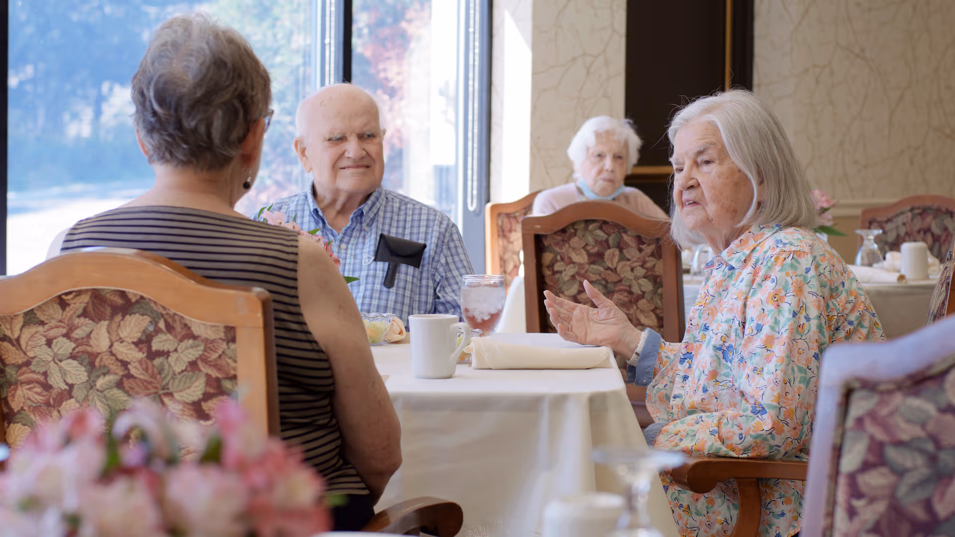 Elderly residents sit and converse around a table in a bright dining room with floral-upholstered chairs.