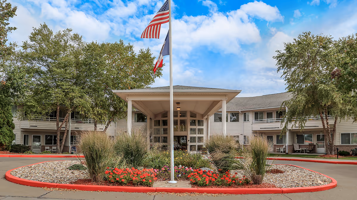 Front exterior view of Holiday Palmer Hills facility with a circular driveway, landscaped garden with flowers and tall grasses, and three flagpoles displaying the American flag and other flags under a partly cloudy sky.
