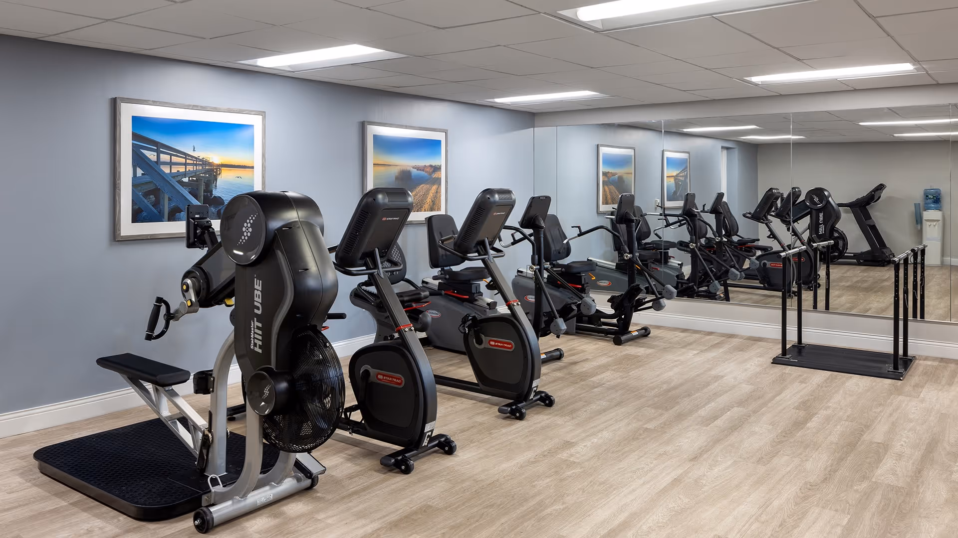 A fitness room with exercise equipment including a HIITUBE machine, two stationary bikes, and a treadmill. The room has light wood flooring, light gray walls, two framed landscape photos, and a large mirror covering one wall.